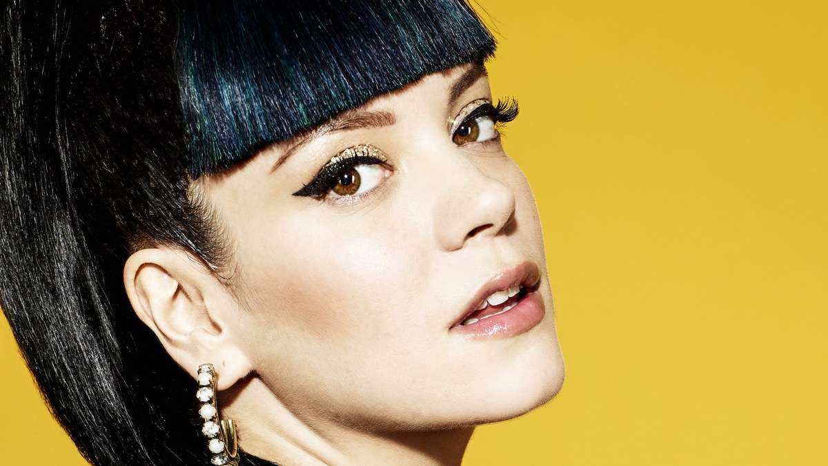 Lily Allen with dark glossy bangs and gold eye makeup looking over her shoulder against a yellow backdrop