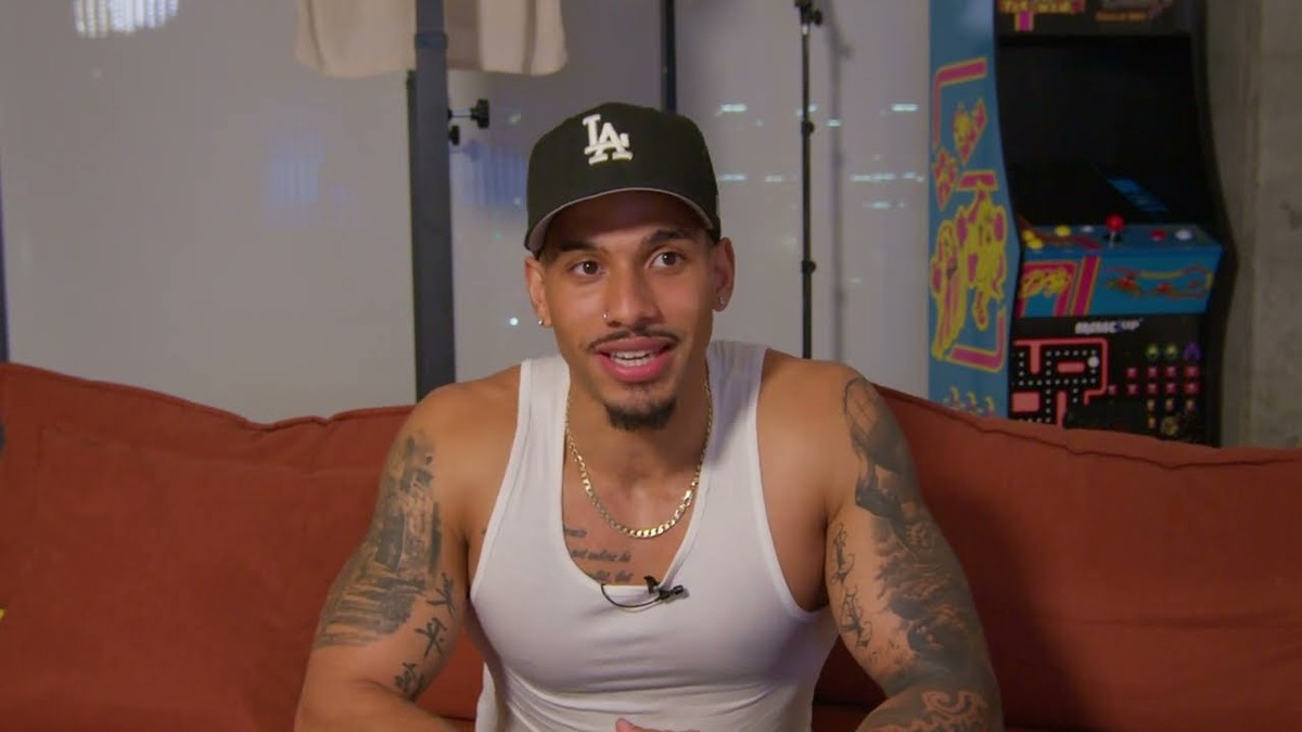 Louis Russell in a black LA cap and white tank top sitting on an orange couch