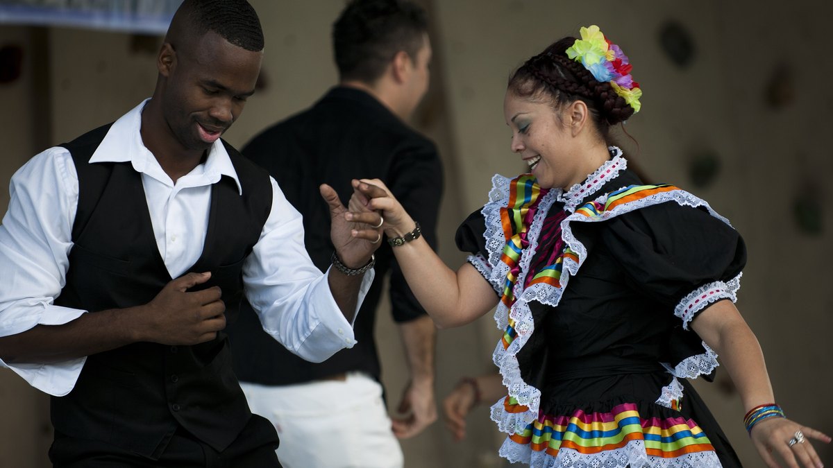 A young woman in a colorful ruffled folk dress dances with a partner in a white shirt and black vest at a Hispanic heritage festival