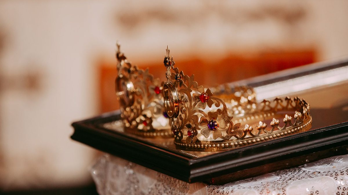 Two ornate gold crowns set with colored stones resting on a dark tray over a white lace cloth