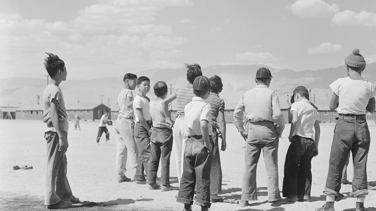 Japanese American boys gathered around a baseball game on dusty ground at the Manzanar camp with barracks and mountains behind them