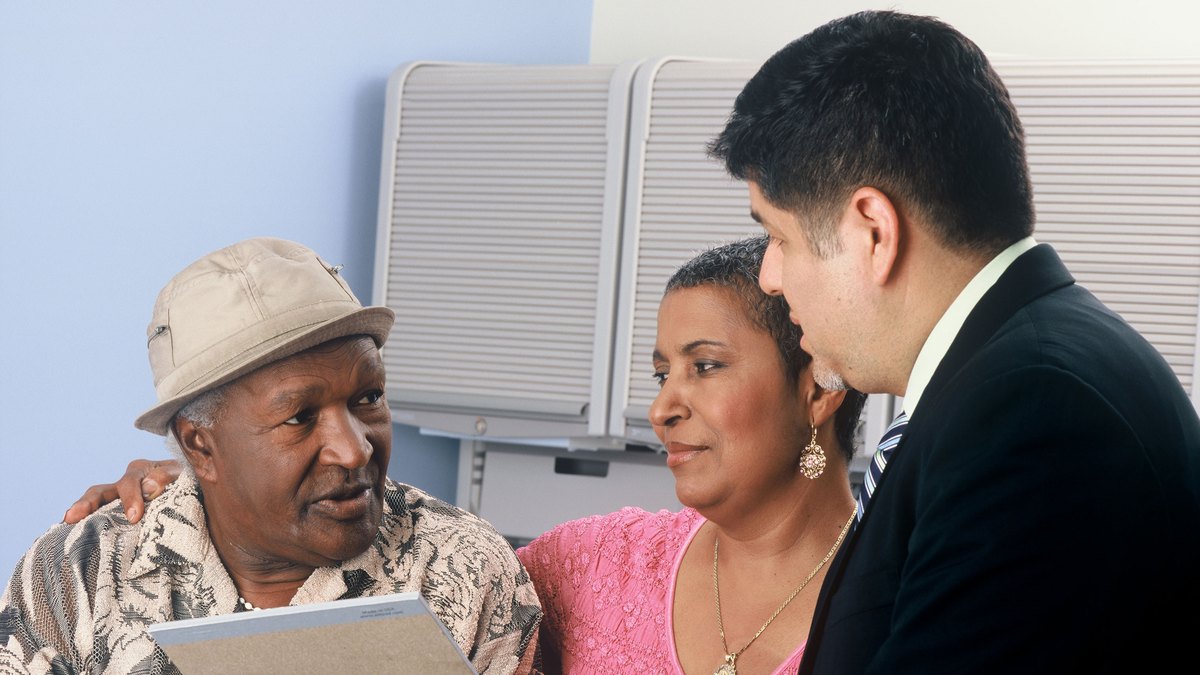An older man in a sun hat and a woman beside him talk over paperwork with an adviser in a suit
