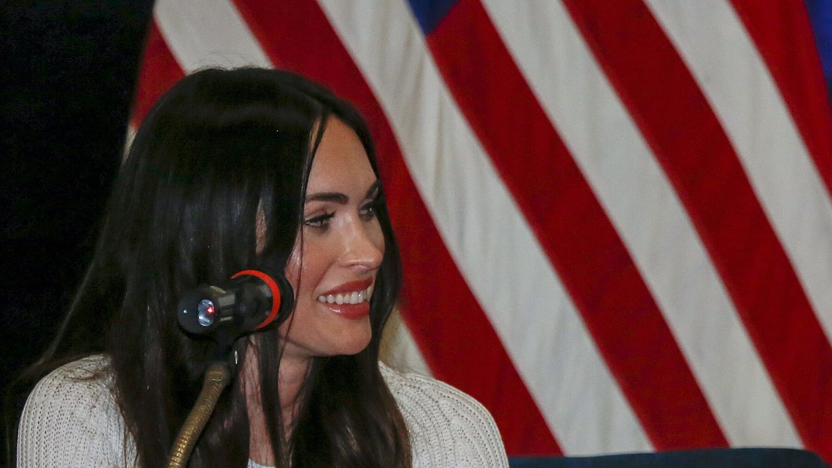 Megan Fox smiling at a microphone in front of a US flag during a 2019 visit with US troops
