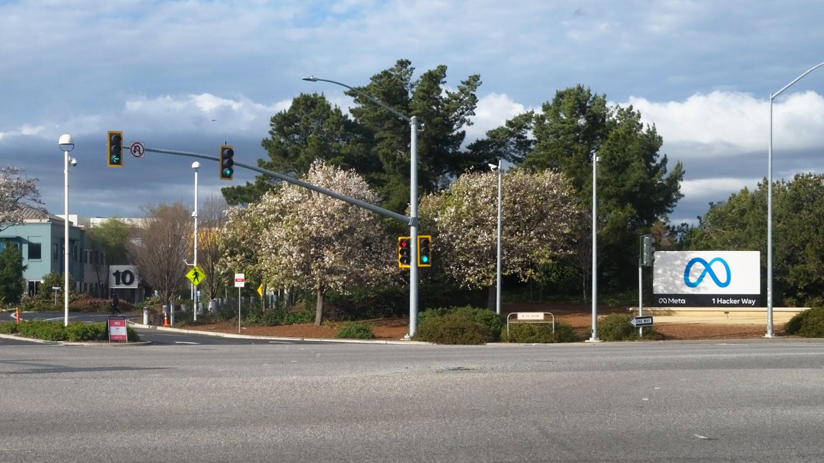 The white Meta sign at the 1 Hacker Way entrance to the company's headquarters in Menlo Park beside a busy intersection with traffic lights and blooming trees