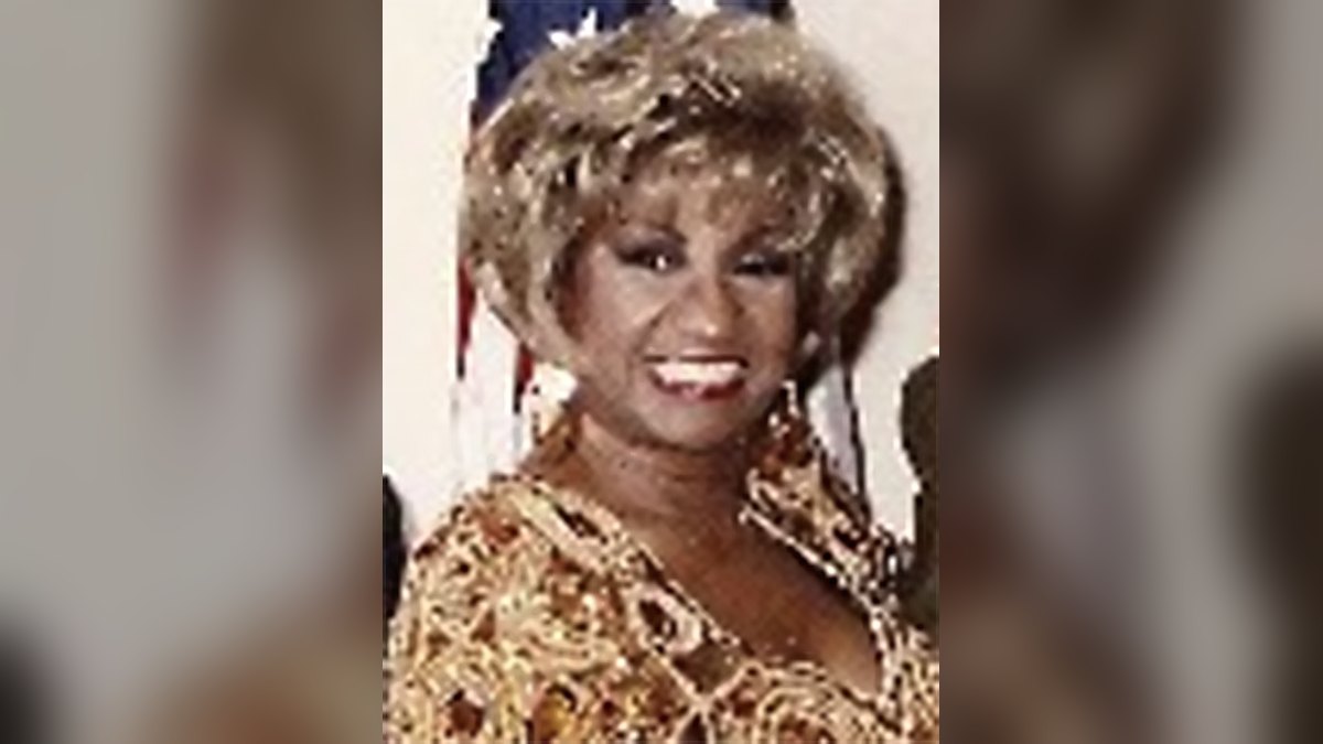 Celia Cruz smiles broadly with her blond curls piled high while wearing a sparkling gold outfit with an American flag behind her
