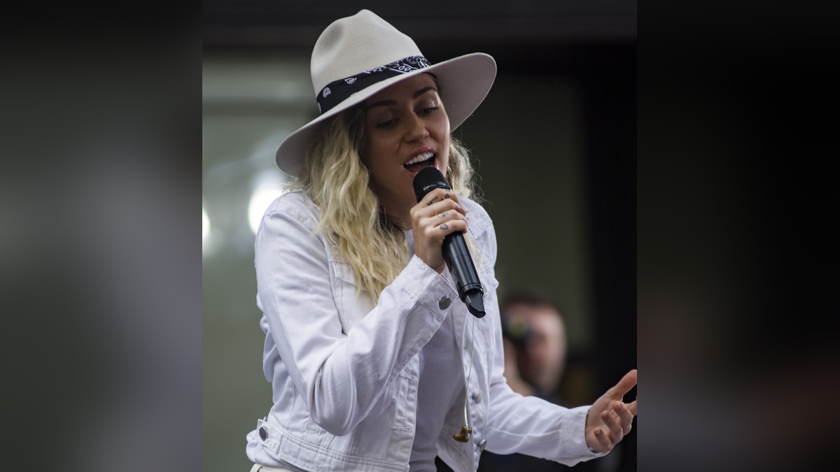 Miley Cyrus singing into a microphone in a white denim jacket and a wide-brimmed cream hat