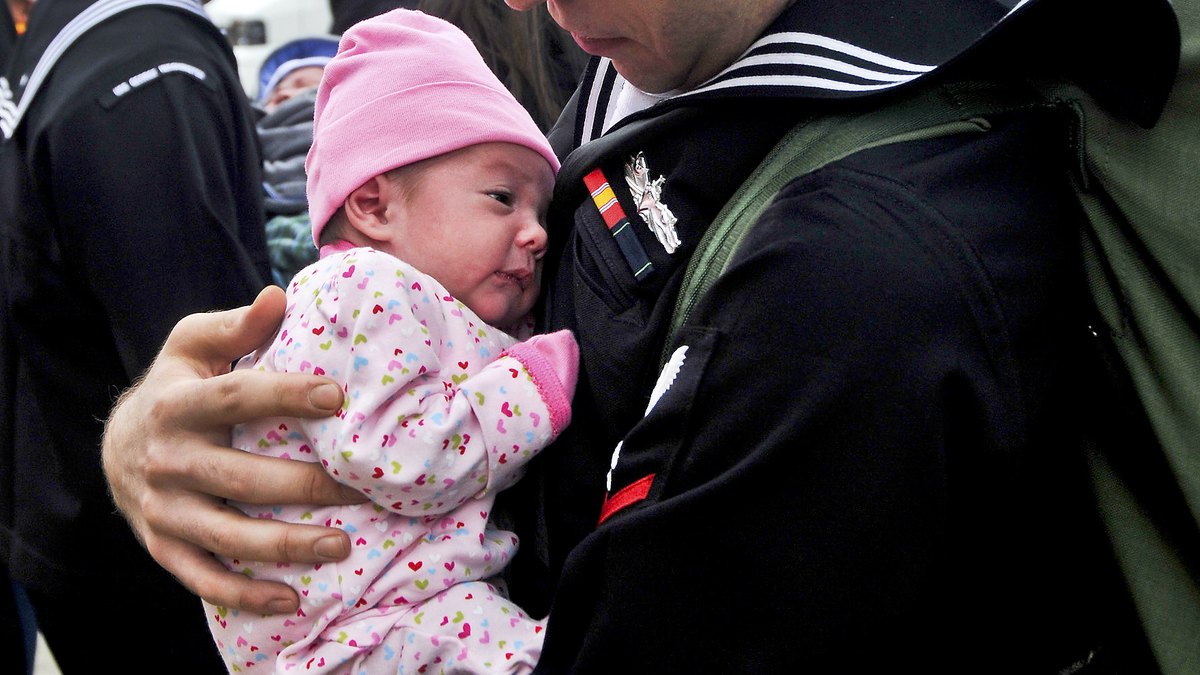 A father in a Navy uniform holds his newborn daughter in a pink knit cap close to his chest