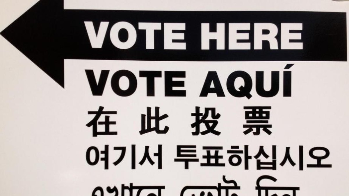 A black and white polling place sign with an arrow reads Vote Here and Vote Aqui above the same message in other languages