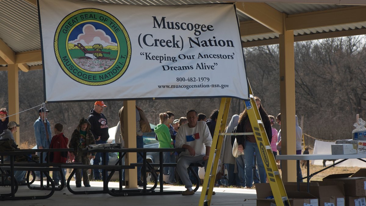 A Muscogee Creek Nation banner hangs under a covered pavilion where people gather at picnic tables