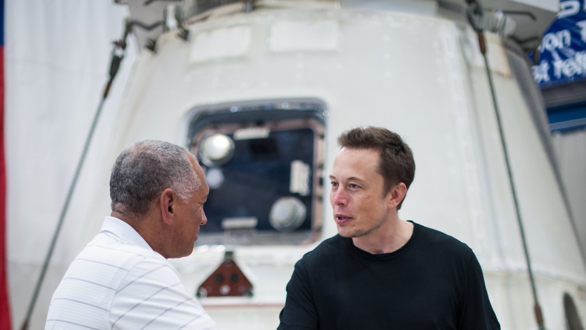 Elon Musk in a black T-shirt talks with then-NASA Administrator Charles Bolden beside a SpaceX Dragon capsule