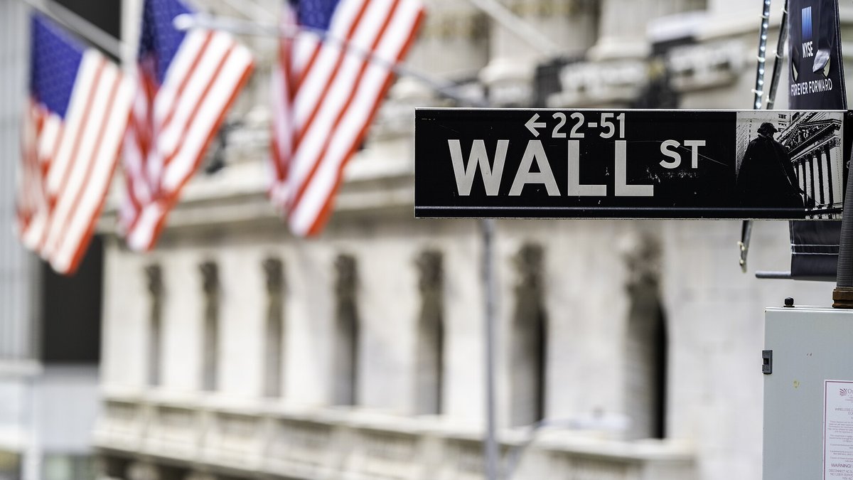 A black Wall Street sign in front of the columns of the New York Stock Exchange with three American flags hanging from the building behind it