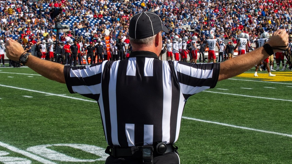 A football referee in a black-and-white striped shirt stands with arms outstretched on the field in front of a packed stadium