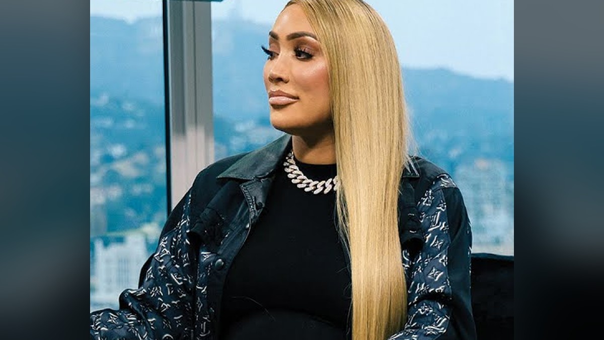 Nikki Mudarris with long straight blonde hair wearing a chunky diamond chain necklace and a patterned black jacket seated in profile in front of a hillside city view