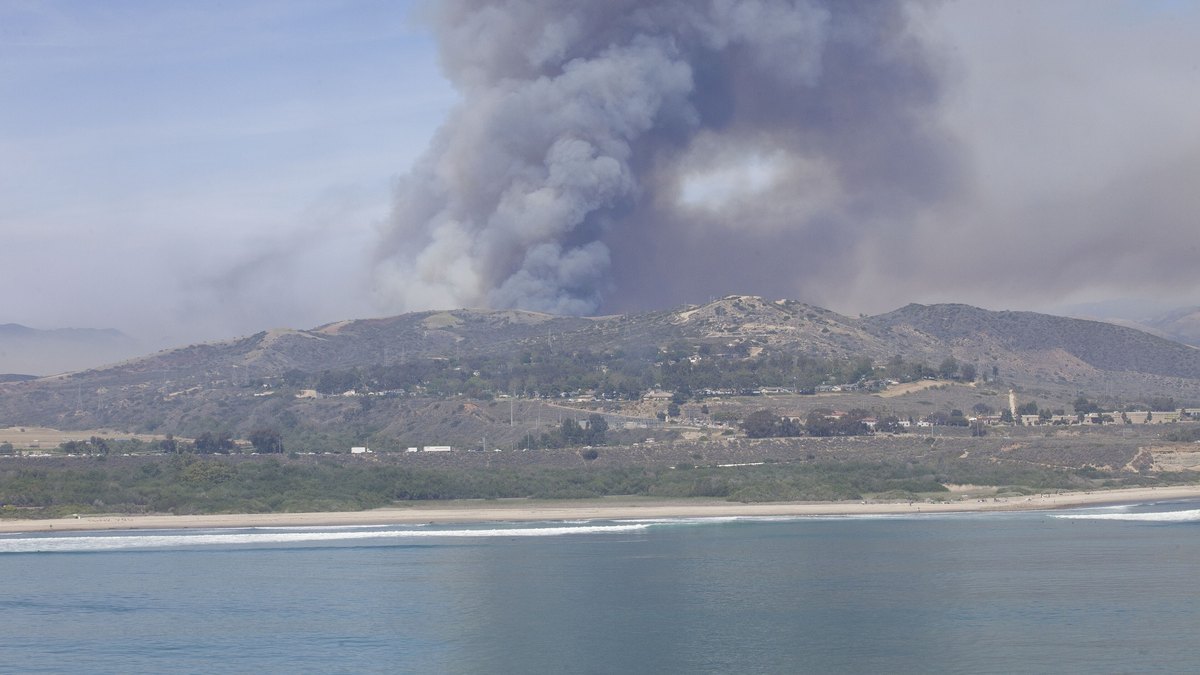 A tall plume of gray smoke rises from a wildfire in the coastal hills of Southern California above the Pacific shoreline
