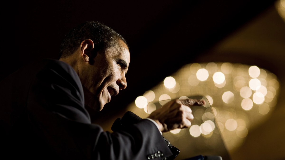 Former President Barack Obama in a dark suit leans over a lectern and gestures as he speaks with warm chandelier lights glowing behind him