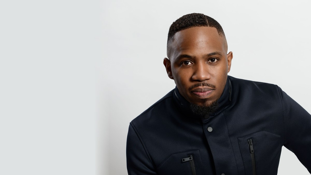 Israel Ford in a dark navy zip-pocket jacket posing against a plain light gray studio backdrop
