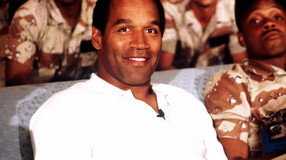O.J. Simpson smiling in a white polo shirt on a couch beside US service members in desert camouflage