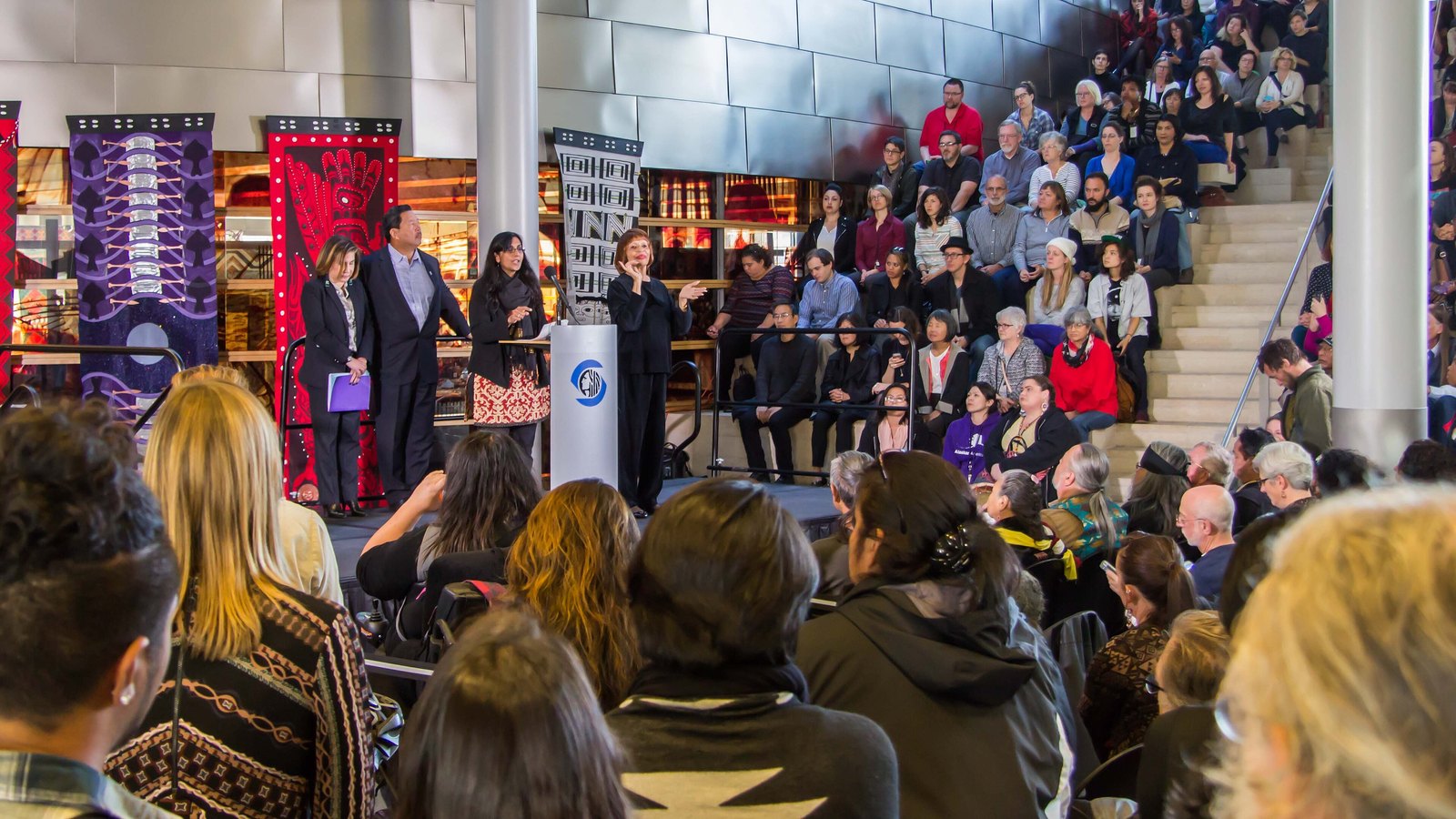 Crowd gathered at an Indigenous Peoples Day celebration as speakers address them from a stage