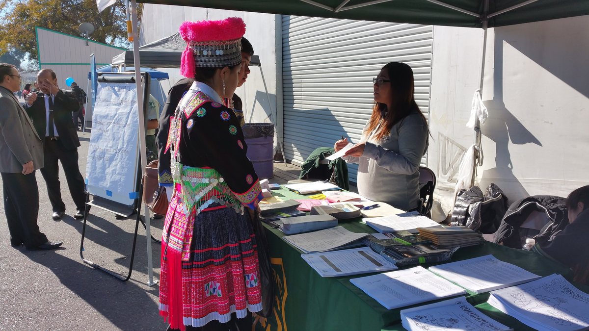 A woman in a traditional Hmong costume with a red hat stands at an information table during a Hmong New Year event