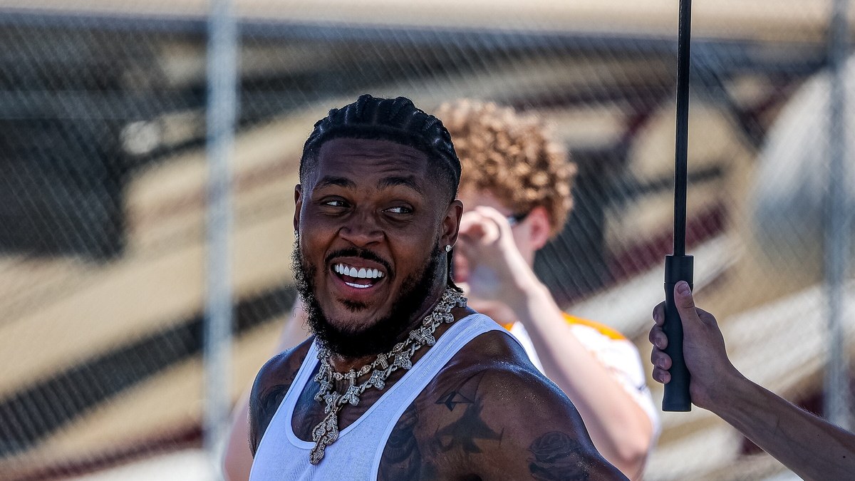 Packers running back Josh Jacobs in a white tank top and a diamond chain smiles and looks over his shoulder at a racetrack in 2025