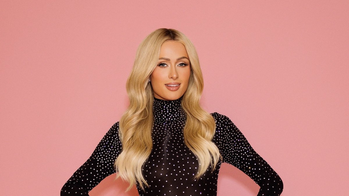 Paris Hilton posing in a sparkly black top against a pink backdrop