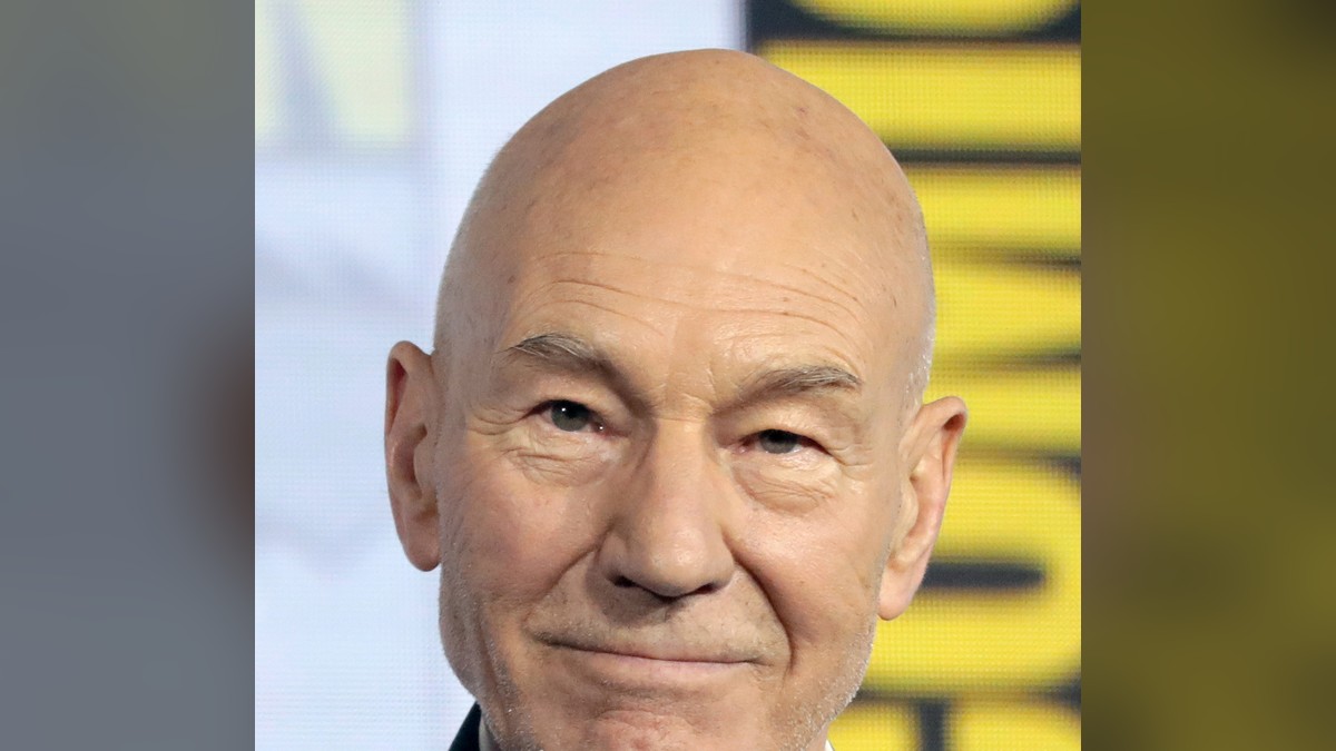 Patrick Stewart smiling warmly in a dark blazer and open white shirt at a fan convention