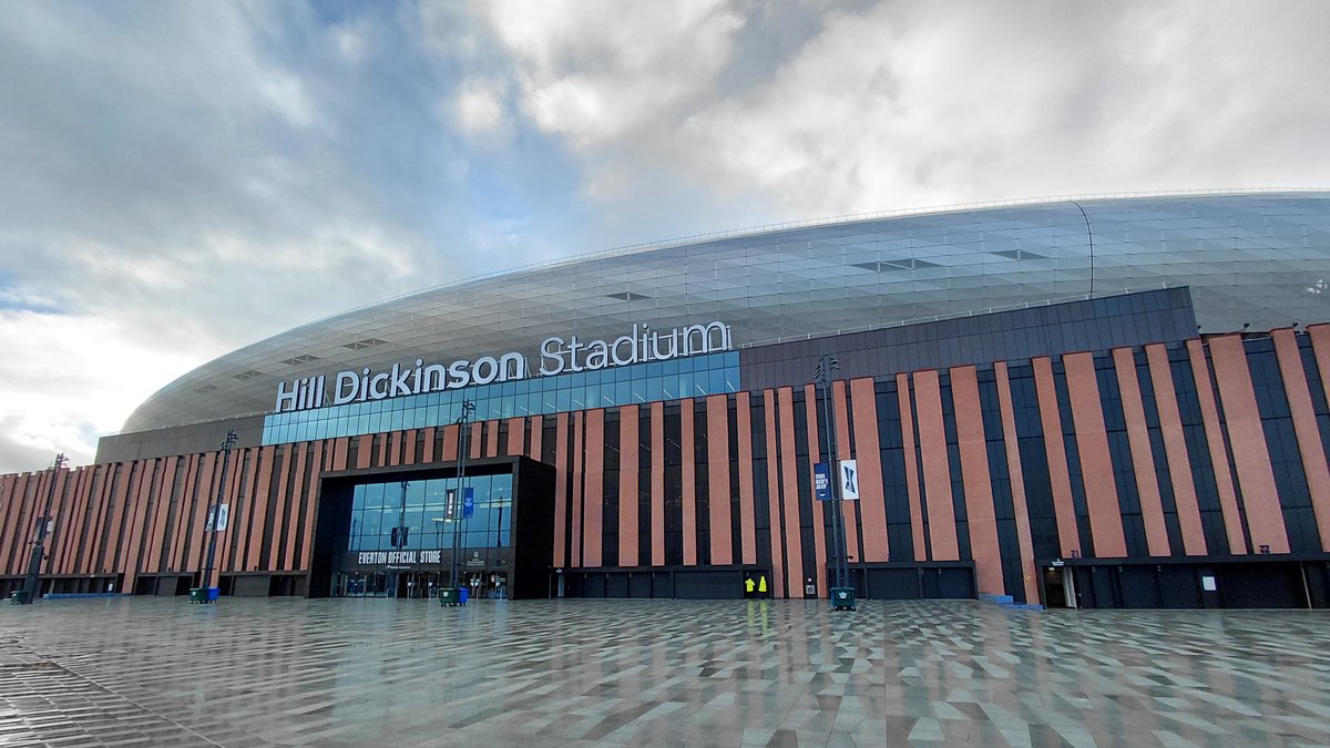 The curved silver roof and lettered sign of Hill Dickinson Stadium in Liverpool rise above a wide wet plaza under a cloudy sky