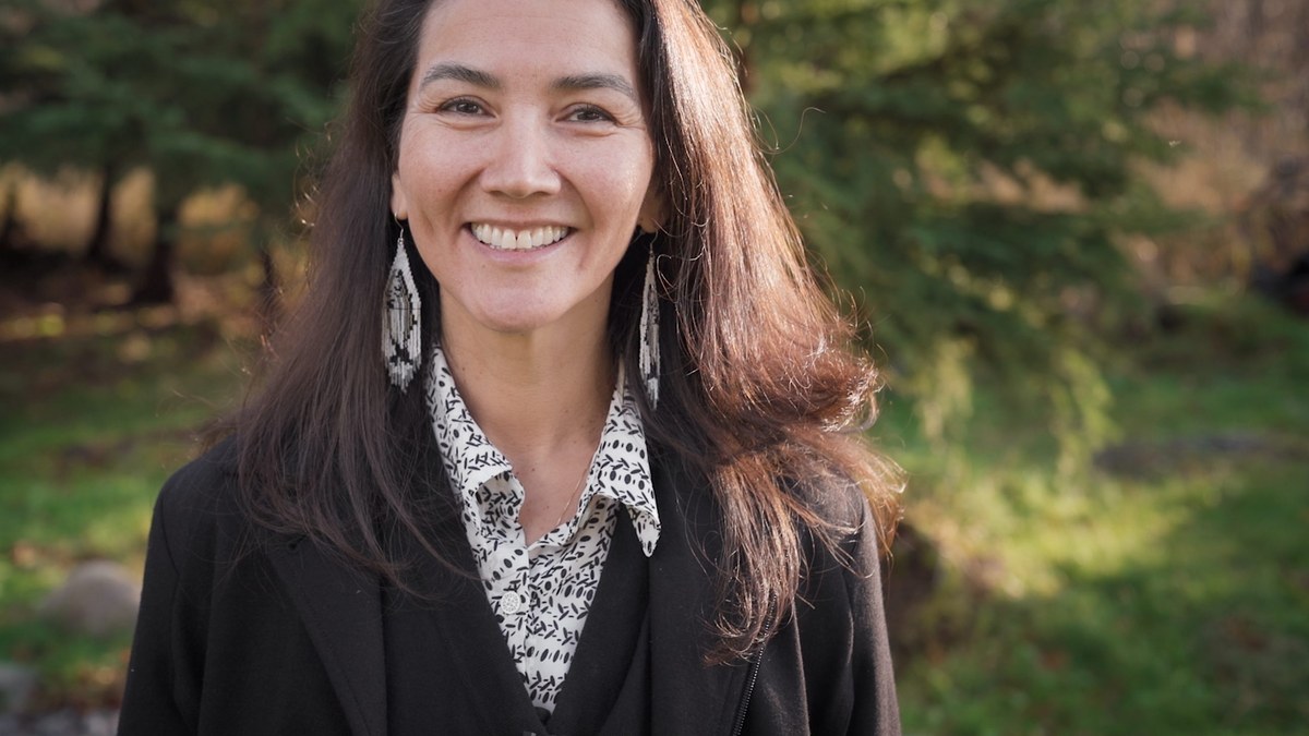 Mary Peltola smiles in a dark jacket and patterned scarf with evergreen trees and green grass behind her