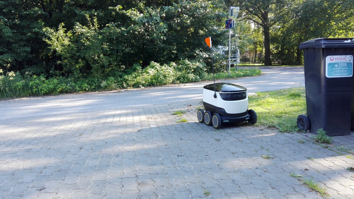 A small white six-wheeled sidewalk delivery robot parked on a brick path near trees