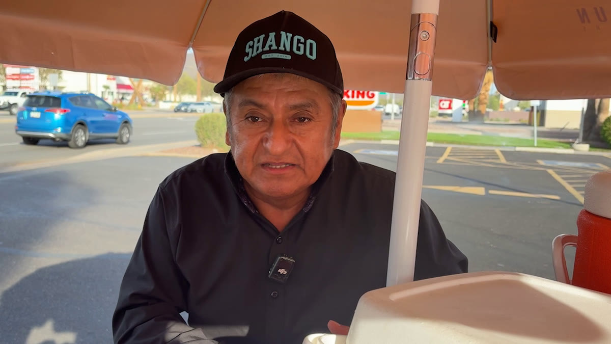 Elote vendor Hilario Cruz Cruz in a black cap and shirt sitting under the umbrella of his food cart in Phoenix