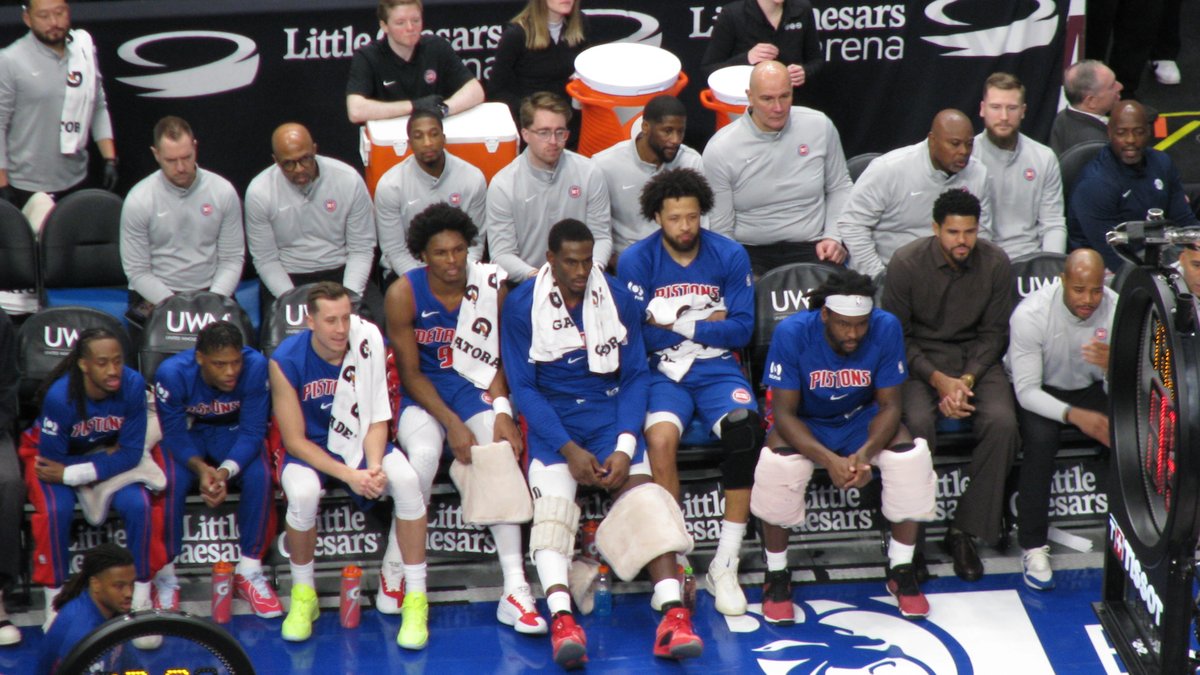 Detroit Pistons players in blue warmups sit together on the team bench during a home game