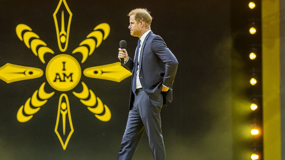 Prince Harry in a navy suit holding a microphone on a stage lit with yellow lights