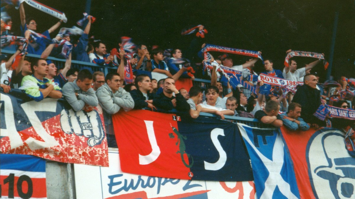 Paris Saint-Germain supporters wave red and blue scarves behind club banners in a stadium stand