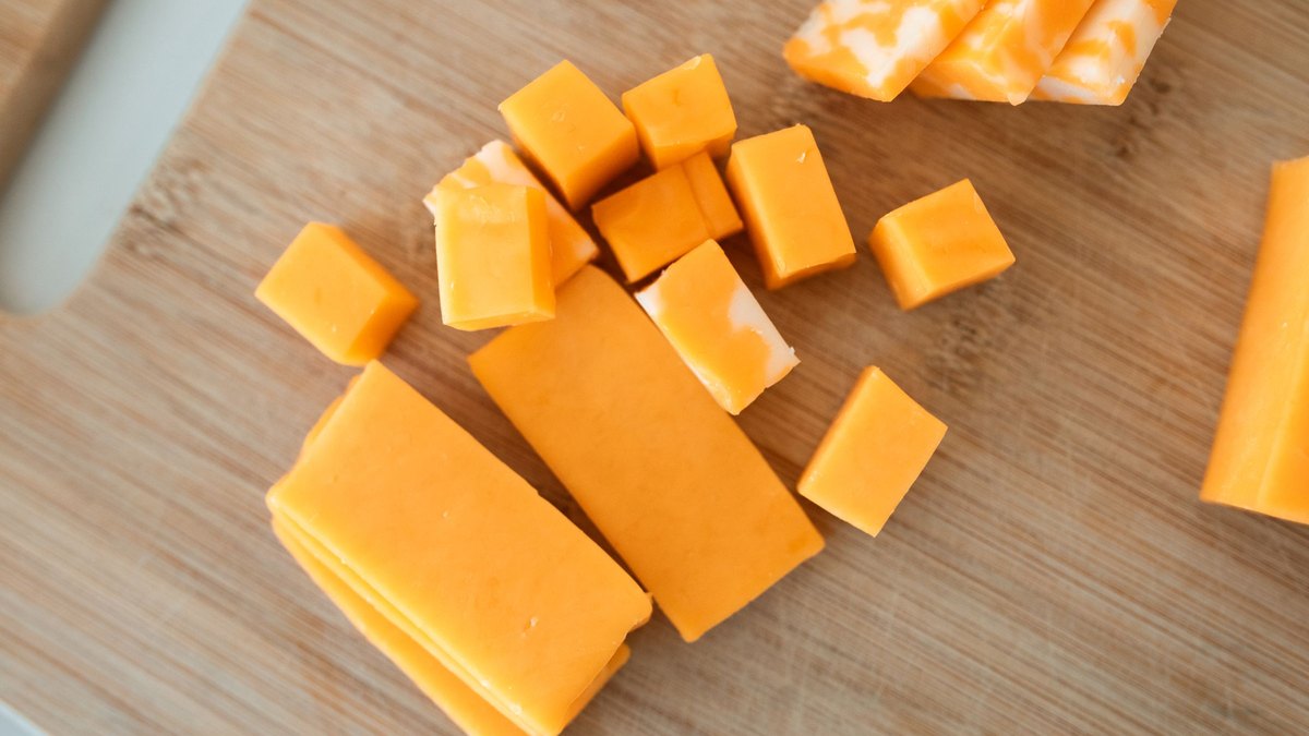 Yellow cubes and blocks of cheddar cheese sit on a wooden cutting board