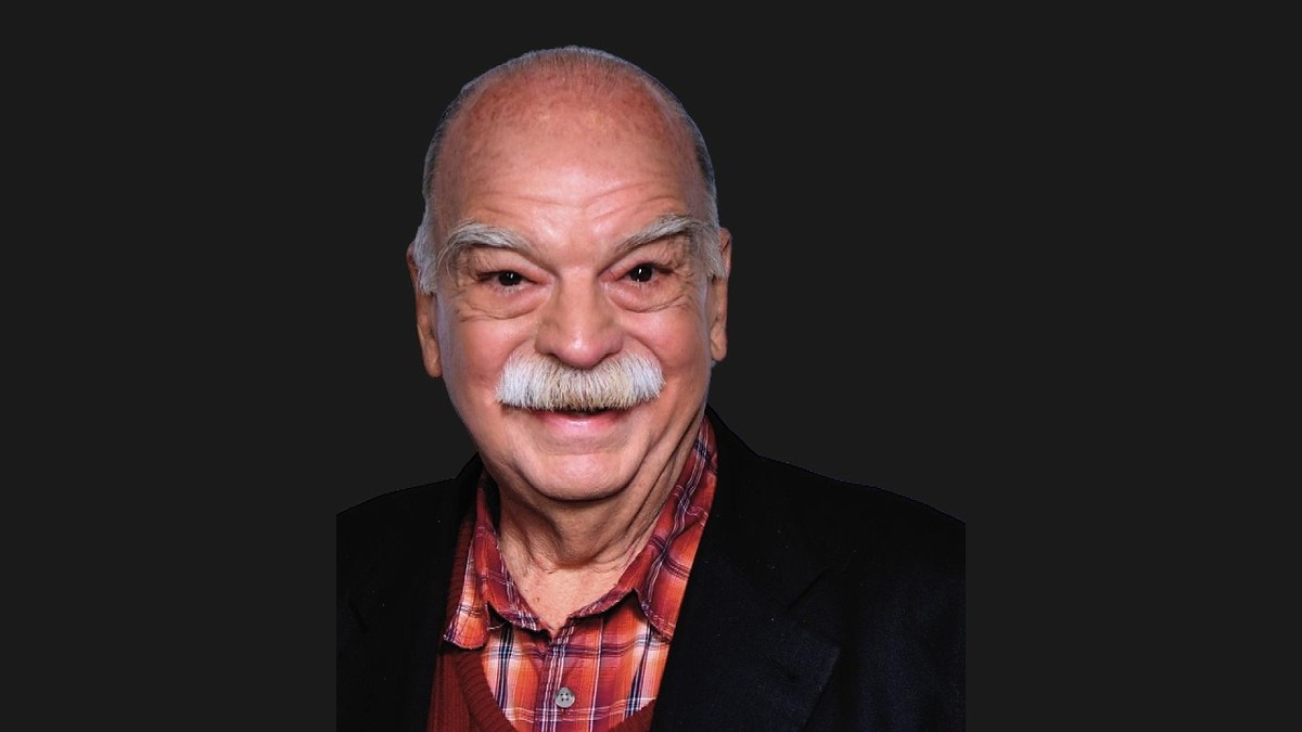 Richard Riehle smiling with a white mustache in a red plaid shirt and dark blazer