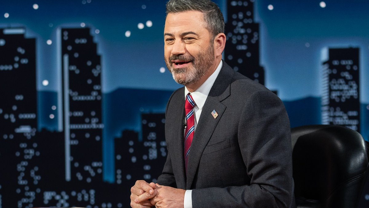 Jimmy Kimmel in a dark suit smiling at his desk on the Jimmy Kimmel Live set