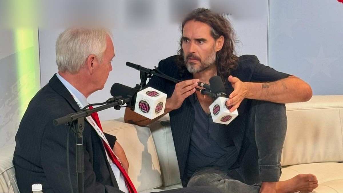 Russell Brand with long hair and a gray beard talking into a microphone during a sit-down interview
