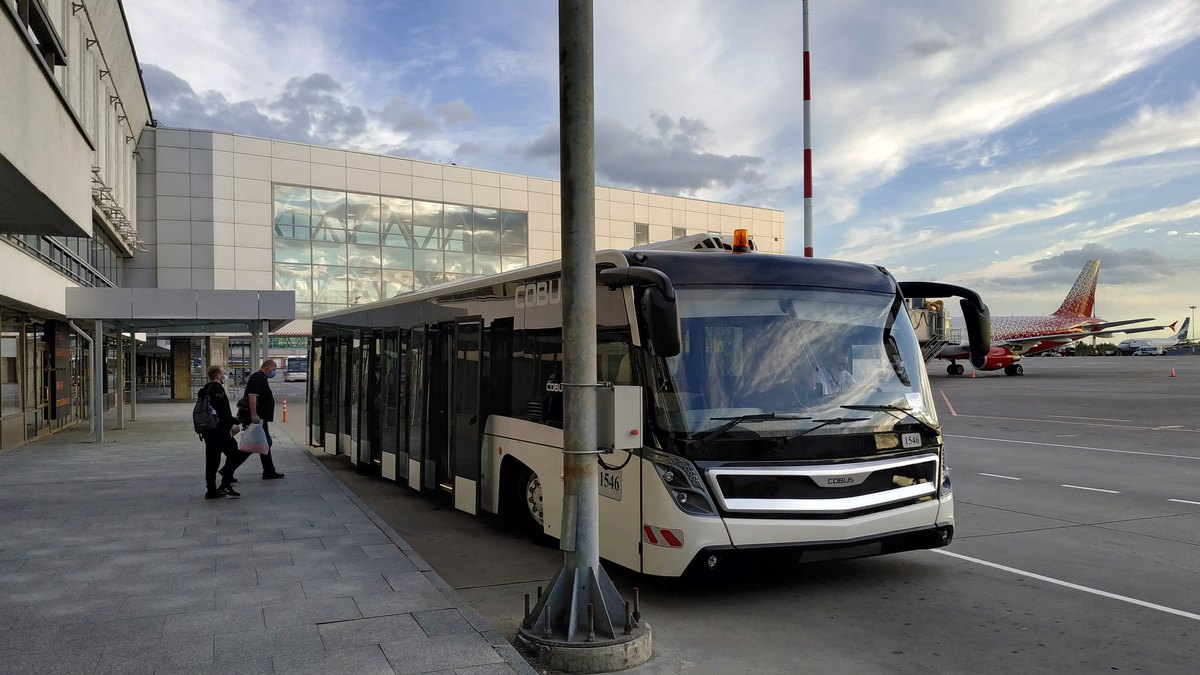 A white airport shuttle bus parked at the curb outside the glass terminal of Pulkovo Airport in St. Petersburg with a jet on the tarmac behind it