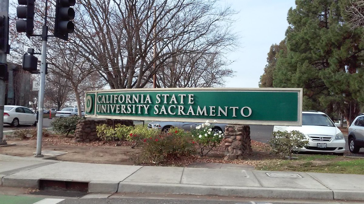 The green California State University, Sacramento sign stands beside a landscaped campus entrance lined with trees