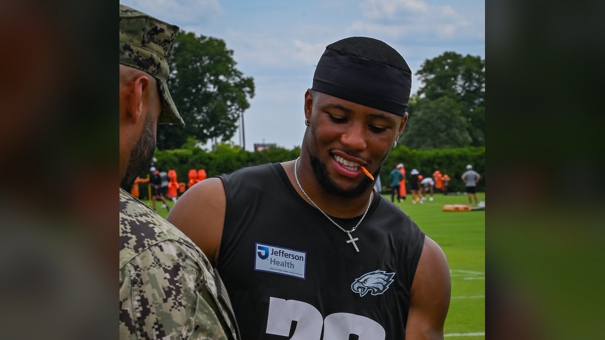 Saquon Barkley smiling in a black Eagles No. 26 practice jersey as he signs a football for a service member at training camp in 2025