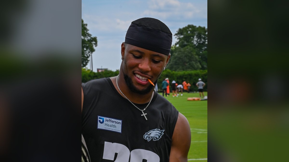Saquon Barkley smiling in a black Philadelphia Eagles practice jersey while holding a football