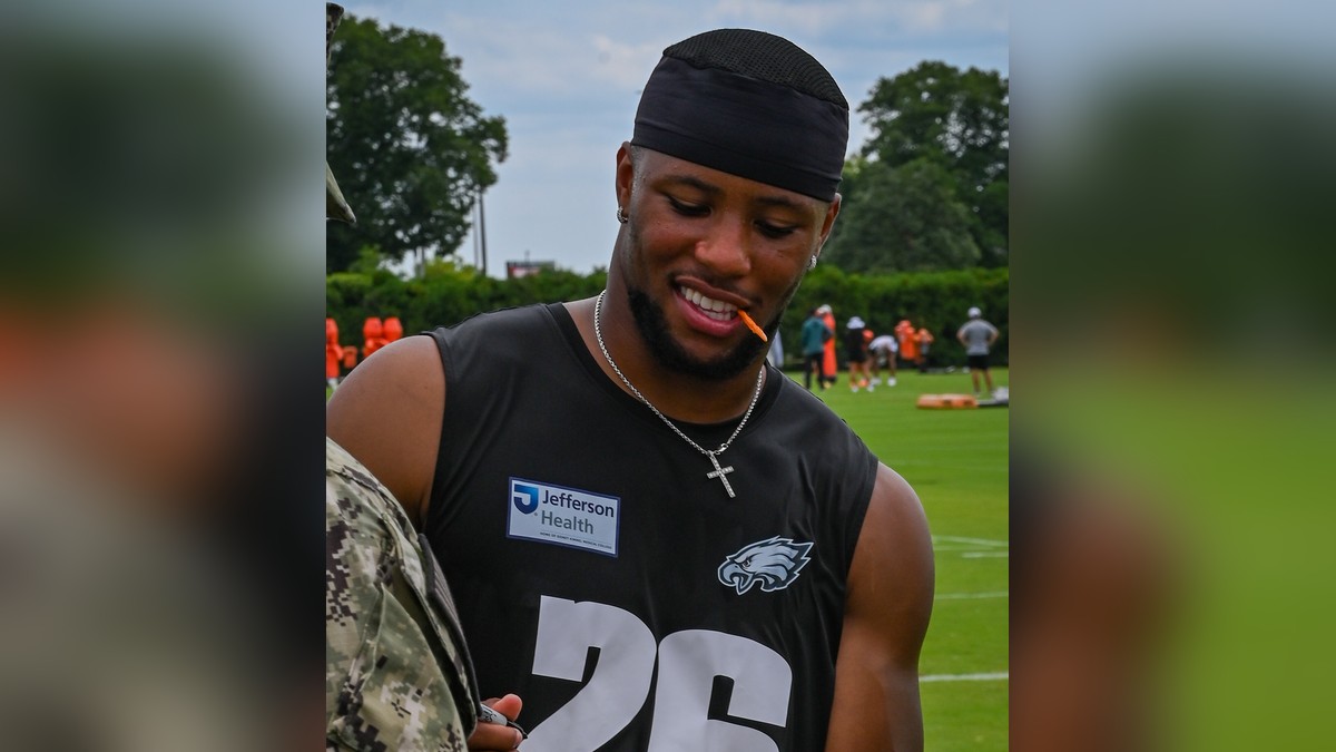 Saquon Barkley smiling in a black Eagles No. 26 practice jersey while signing a football for a service member at training camp