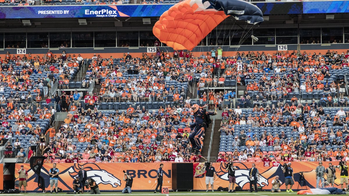 A skydiver under an orange and navy canopy descends toward the field at the Broncos stadium in Denver as fans in orange fill the stands