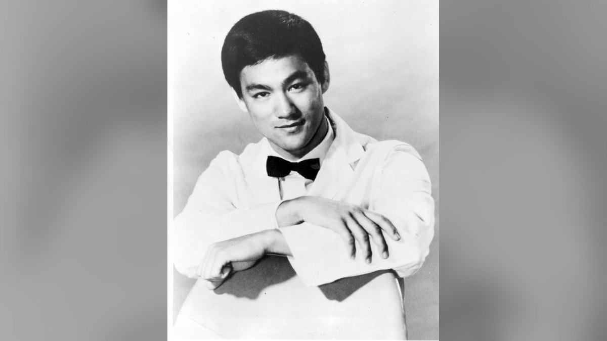 Black and white 1967 portrait of Bruce Lee in a white jacket and bow tie with his arms folded