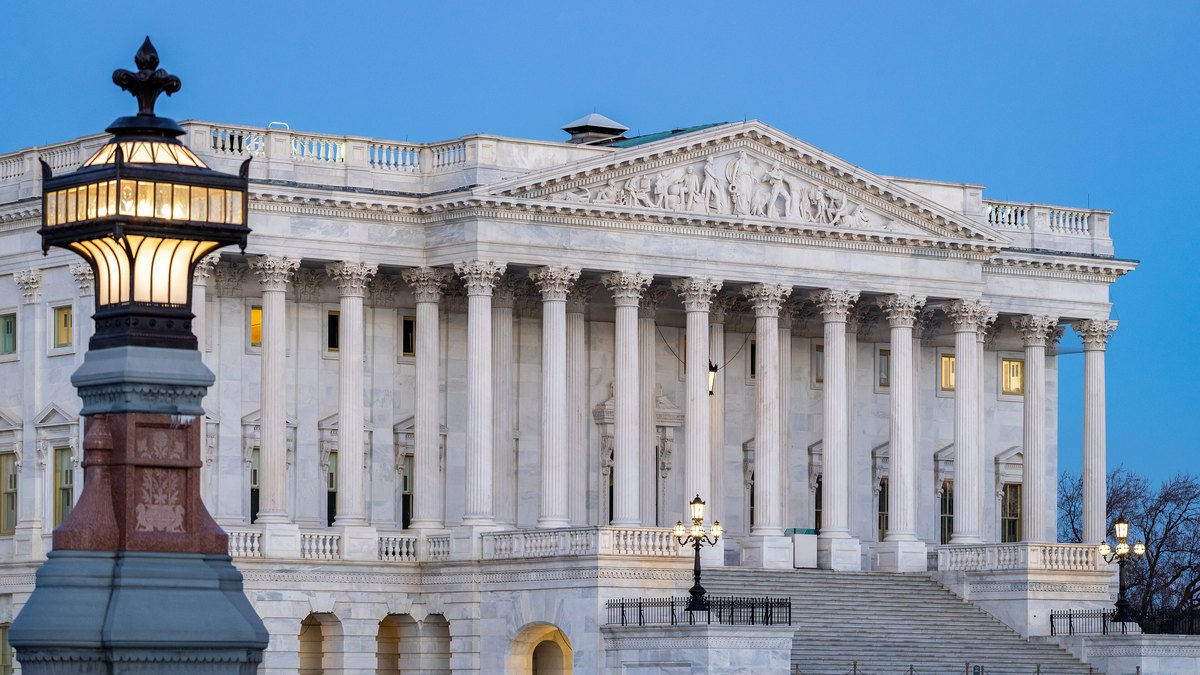 The white marble Senate wing of the U.S. Capitol at dusk with lit lamps and wide stone steps under a deep blue sky