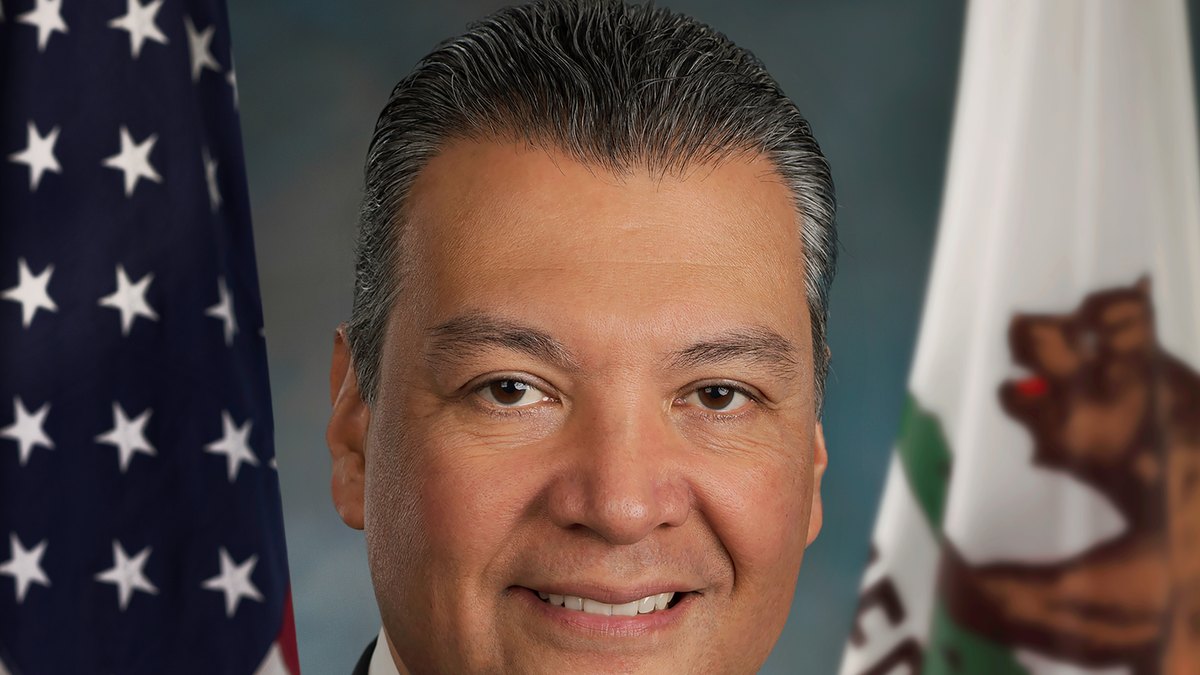 Sen. Alex Padilla smiling in a dark suit and striped tie in front of the American and California flags