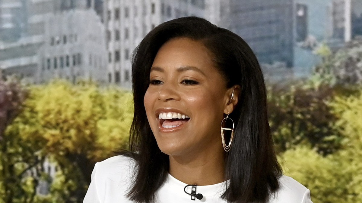 Sheinelle Jones laughing at a morning show desk with a city skyline and trees behind her