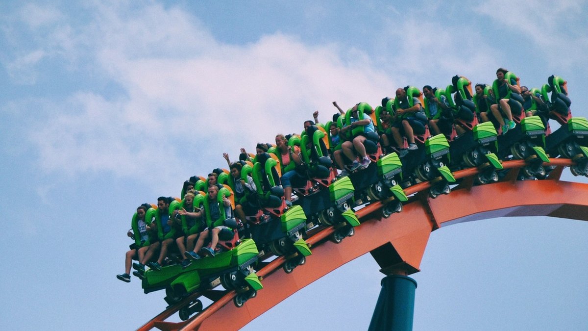 A roller coaster train climbing an orange steel track against a pale blue sky