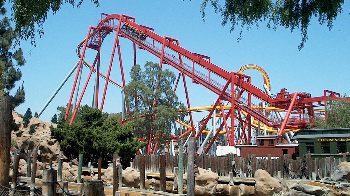 A red steel roller coaster lift hill rising steeply under a clear blue sky