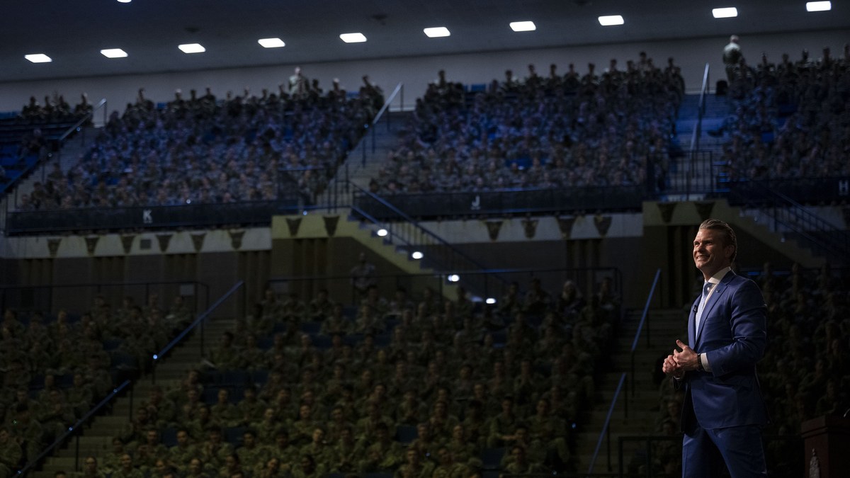 Pete Hegseth smiling on a stage with his hands clasped in front of a large crowd of uniformed midshipmen in an arena